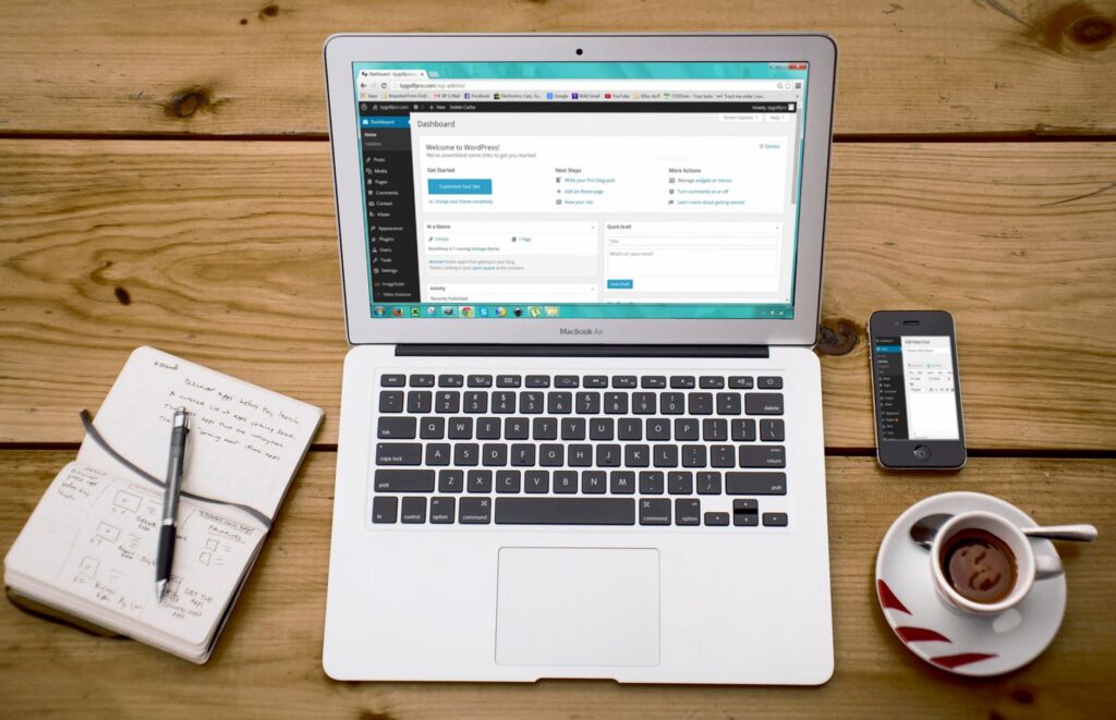 Laptop showing the WordPress dashboard on a wooden desk alongside a notebook and coffee, representing the manual WordPress publishing workflow