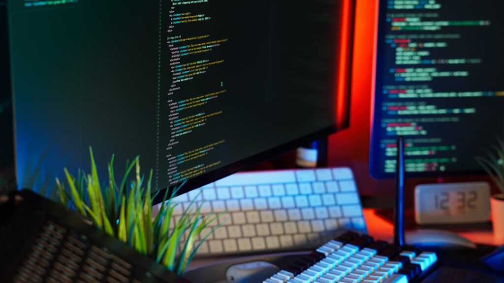 Dual-monitor developer workstation showing code on screens with colorful syntax highlighting, keyboard, and ambient RGB lighting.