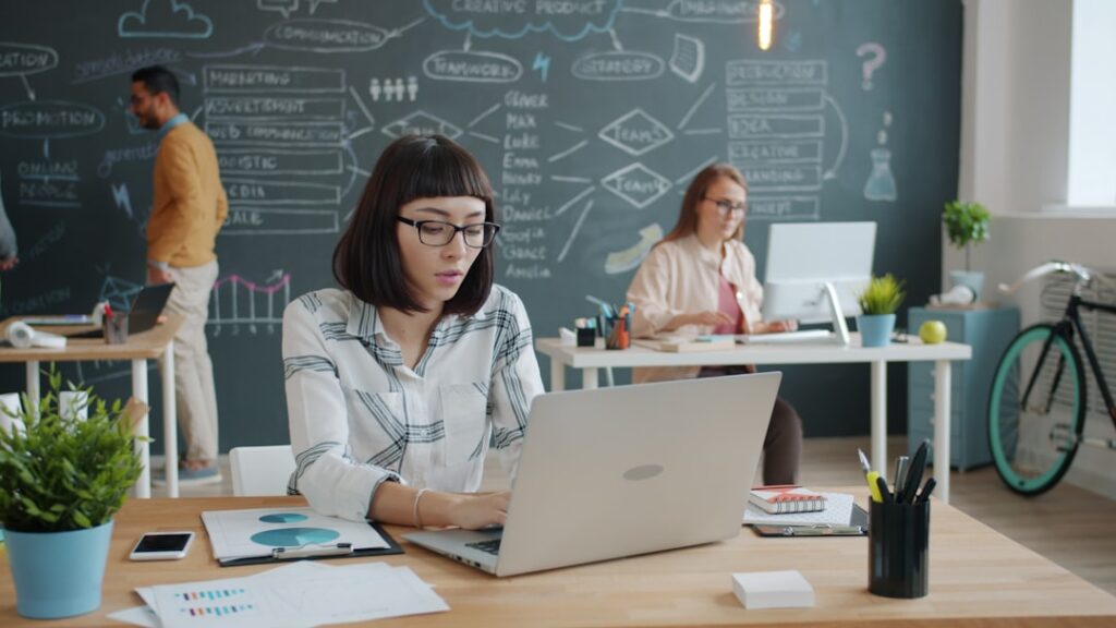 A content strategist working at a laptop in a creative office, with a strategy diagram visible on a chalkboard wall behind her, representing the human review layer that automation cannot replace