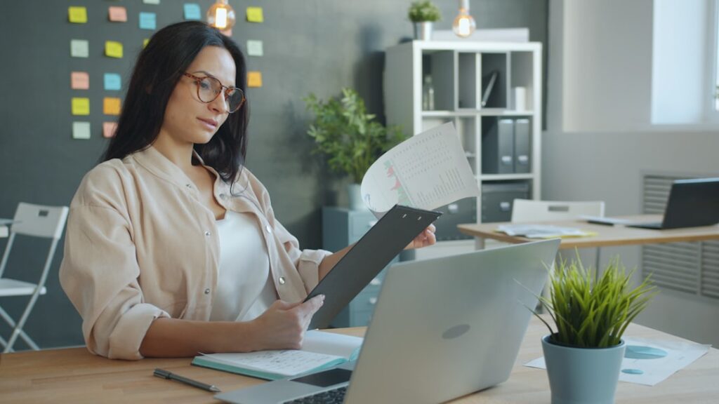 Businesswoman reviewing a document alongside a laptop screen, representing the human approval step in a content automation workflow.