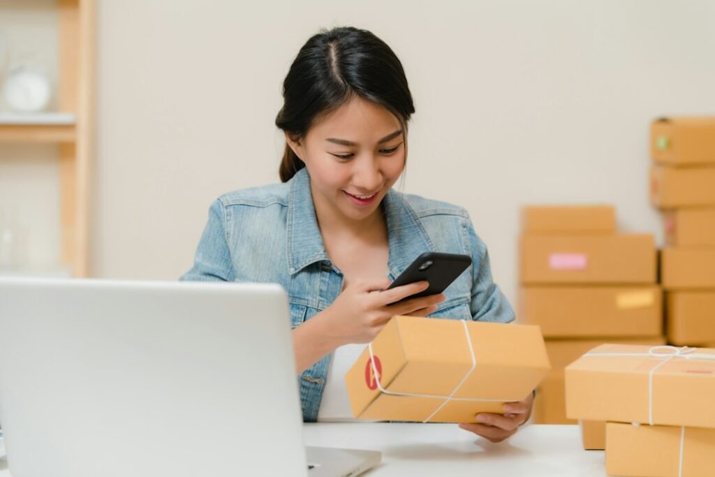 Small business ecommerce seller scanning product barcodes with smartphone surrounded by shipping boxes and a laptop
